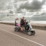 Scooterpac Tandem two-person mobility scooter side view with elderly couple riding by seaside